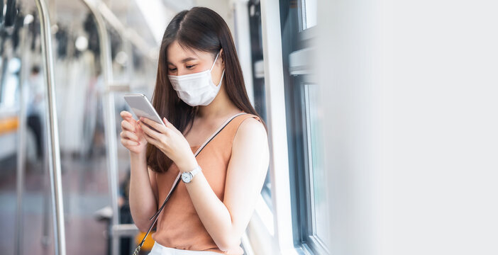Young Asian woman passenger wearing surgical mask and listening music via smart mobile phone in subway train when traveling in big city at Covid19 outbreak, Infection and Pandemic concept