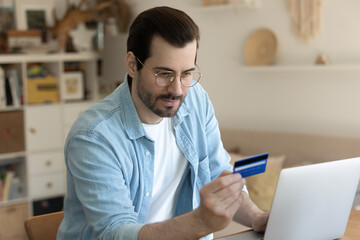 Young Caucasian man in glasses make online payment or purchase on laptop with credit card from home, millennial male shopping on internet or paying bills using web banking service system on computer