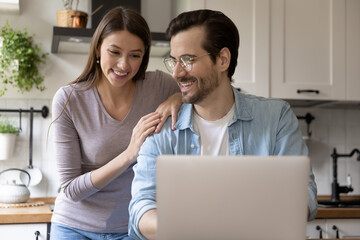 Happy young Caucasian couple sit in kitchen look at laptop screen browsing internet surfing web together, smiling millennial husband and wife shopping online on computer at home, use gadget technology