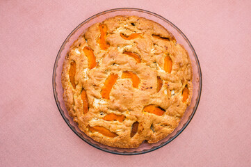 A large, round baked apple and apricot pie stands in a glass form on a table with a pink tablecloth. Delicious, fresh dessert.