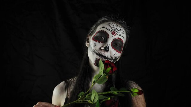 Halloween mask Calavera Catrina. Girl with a rose in her hands. Mexican day of the dead. Portrait of a young woman with a terrible multi-colored makeup for Halloween on a dark background. 4K. 