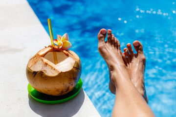 Fresh coconut with a glass straw decorated with Plumeria flower. Woman's feet near the swimming pool. Tropical mood. Bali, Indonesia.
