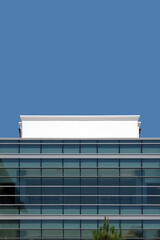 Fototapeta premium Low angle view of a section of a modern building with a glass window facade and a white concrete top part under blue sky