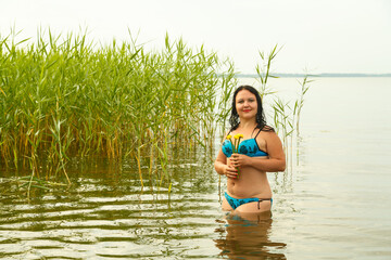 A brunette woman in a blue swimsuit at dawn in the azure water in the reeds with water flowers.