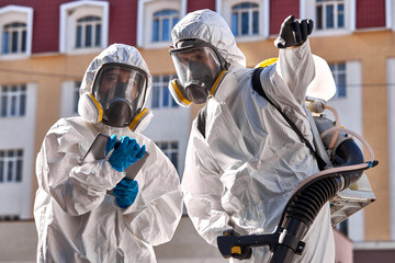 professional specialized fearless team in white protective suits and masks ready to work, clean streets of big city. two friendly workers discuss dangerous areas for cleaning