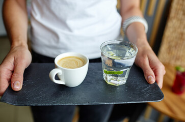 Female hands holding stone plate with cup of coffee and water in a glass with lime.Coffee break.Blurred image,selective focus