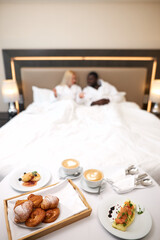 diverse man and woman going to have breakfast on bed, african man and caucasian woman get tasty food by hotel staff, they enjoy wekeends together, lie on bed