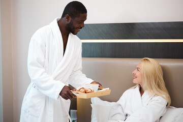 young african man treats his caucasian wife with breakfast, love her. happy couple at weekends in hotel, morning