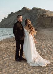 An elegant bride with disheveled curls in a sparkling dress with a large hemline stands on the sand...