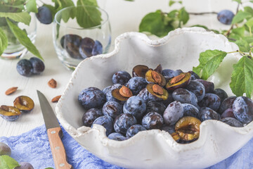 White curved bowl filled with ripe plums on a white wooden table with green leaves, some of them sliced. Horizontal closeup.