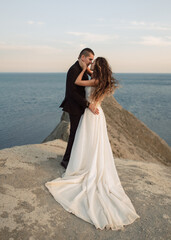 Fototapeta na wymiar Sensual and beautiful wedding couple passionately embraces on top of the cliff overlooking the sea. A bride in a fashion wedding dress with a chic hem snuggles up to her beloved.