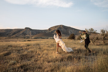 Bride and groom run across picturesque field against sunset mountains. Bride with nude legs runs across the autumn field against mountains in the safari park. Luxury wedding dress flutters and slides © Damien
