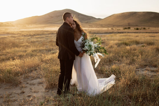 Wedding couple embraces in the Safari on the background and mountains in sunlight. Beautiful bride closed her eyes and dreams in fashionable wedding dress with luxury bouquet with white peonies.