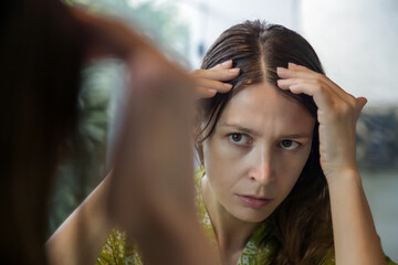 Portrait of a beautiful young woman examining her scalp and hair in front of the mirror, hair roots, color, grey hair, hair loss or dry scalp problem	