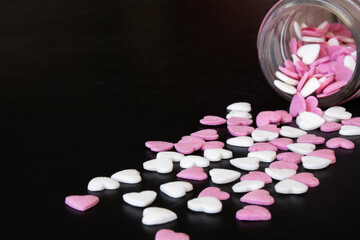 Scattered pink and white small heart-shaped sugar candies falling from glass container on black table background. Empty space for text