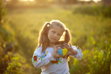 Little girl dressed in an embroidered shirt (vyshyvanka) with a yellow and blue heart in her hands in a  field at sunset. Ukraine's Independence Day. Little patriot