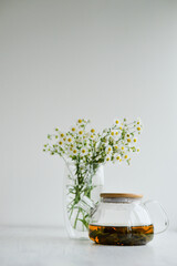 Green tean in glass pot and cup standing on the white background with camomile bouquet