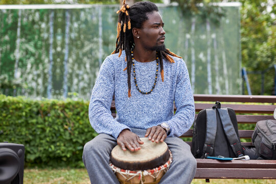 handsome young afro man with djembe drums outdoors. african american man with dreadlocks sits on bench, perform music