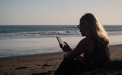 A white girl looks at her phone while sitting on a deserted beach.