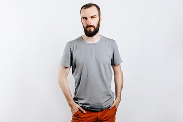 A handsome brutal brunette man with a beard in a gray shirt looks seriously at the camera, holds his hand in his pockets. Place for advertising on a gray background.