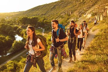 Young people with backpacks walking along narrow path in mountains. Group of cheerful tourists hiking outside in summer - obrazy, fototapety, plakaty