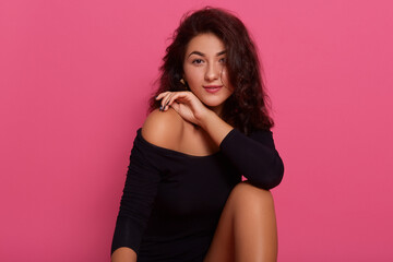 Picture of charming female with pleasant appearance, looking at camera, wearing black bodysuit, posing isolated over pink background, keeping hand near bare shoulders.