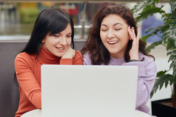 Two women working together, beautiful colleagues working on project while sitting in front of lap top and laughing, ladies have video call in cafe.