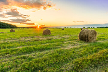 Scenic view at picturesque sunset in a green shiny field with hay stacks, bright cloudy sky , trees and golden sun rays with glow, summer valley landscape