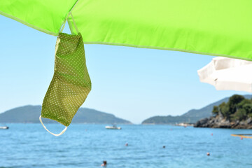 A face shield hanging from a sun umbrella on the beach by the sea. The concept of a beach holiday at sea under the new rules, in connection with the COVID-2019 coronavirus epidemic. Selective focus.