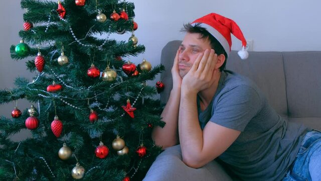 Sad man in red cap is boring sitting on couch near Christmas tree at home. Single young guy in depression on winter holidays. Alone in New Year night. Loneliness and isolation in quarantine pandemic.