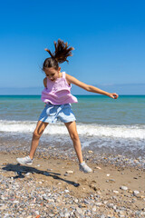 Little girl dressed in pastel colors jumping and playing on the stone and sandy shore of the Mediterranean Sea