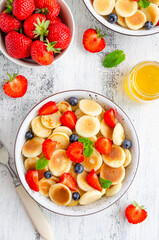 Pancake cereal, mini pancakes in a bowl with fresh strawberries, blueberries and honey. Trend food. Vertical, top view, copy space.