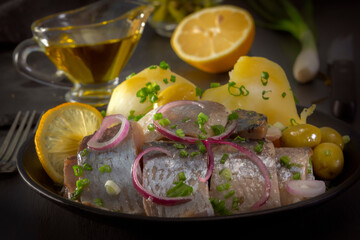 Pickled herring slices with red onion, fermented cucumber, potatoes, olives and lemon in a black plate on a dark wooden table. Close-up