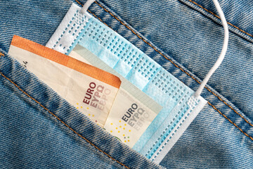 Close-up of a protective medical mask and money in the pocket of old jeans on a wooden background