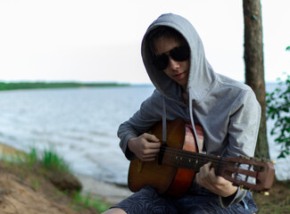 A teenager plays a guitar while sitting on the shore of a lake.