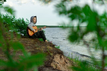 A teenager plays a guitar while sitting on the shore of a lake.