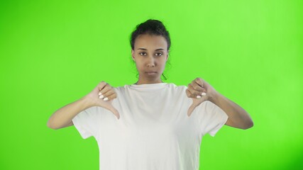 Beautiful young girl in a white T-shirt shows thumb down with both hands on chroma key background