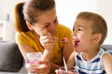 Mother and soon feeding at home in day - Caucasian woman sitting on sofa feed small boy with desert...