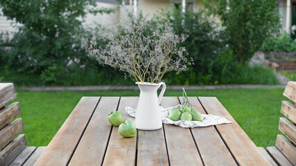 A white jug with a bouquet of wildflowers on a wooden table. Summer still life on the background of the cottage plot. Still life with green tomatoes.