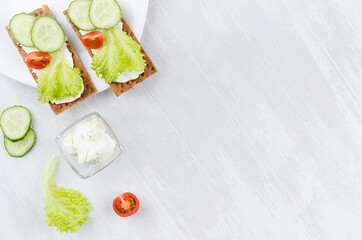 Fresh spring vitamine sandwiches of flat cereal rye dry crisps bread with green lettuce, cucumber, tomato, cream cheese on white wooden table, top view, copy space.