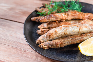 Appetizing fried capelin on a black plate stands on a wooden surface. Simple rustic food concept