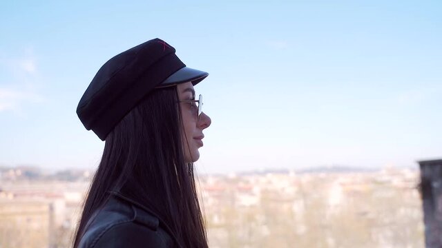 Silhouette of a young stylish girl on a background of sky, light blue background. Portrait of brunette female in sunglasses hat and black leather jacket. Woman fashion model in black clothes