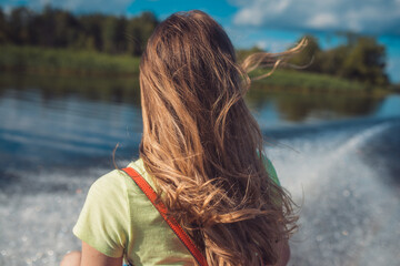 a young woman with long hair rides a fast boat on the water surface of the lake. Back view. Against the background of a white foam trail from the propeller and green shores receding into the distance
