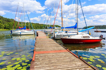 Fototapeta premium Yacht boats and wooden pier on lake shore in Karwica village port on sunny summer day with with clouds on blue sky, Mazury Lake District, Poland
