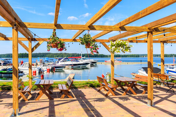 Restaurant tables on terrace with flowers and view of sailing boats on lake Beldany in Piaski port on summer sunny day, Mazury Lake District, Poland