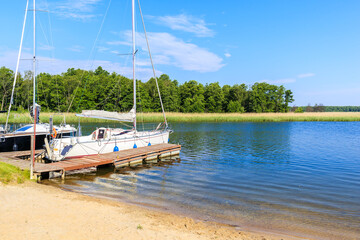 Fototapeta na wymiar Beach in Pisz sailing port on Lake Ros on summer sunny day with yacht boat at pier in background, Masurian Lakes, Poland