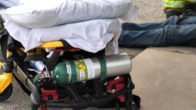 First Responders EMT, Police, And Fire Fighters Helping A Patient Wearing Blue Jeans On The Ground After An Incident Or Car Crash. Focus Is On The Gurney (stretcher) And Oxygen Tank. Shot In 4K 60fps.