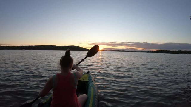 LD Wide shot of a young woman paddling a kayak at sea as the sun is setting behind the coastline in front of her