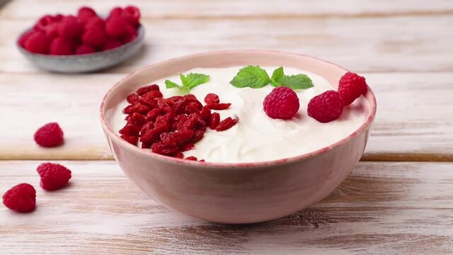 Male hand decorates yogurt with raspberry and goji in ceramic bowl on white wooden background. Side view, close up.