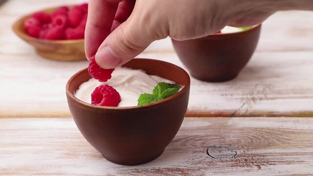 Male hand decorates yogurt with raspberry in clay cups on white wooden background. Side view, close up.